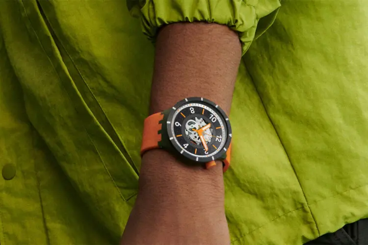 stor swatch klocka med svart boett och orange armband på handled på en person med grön skjorta